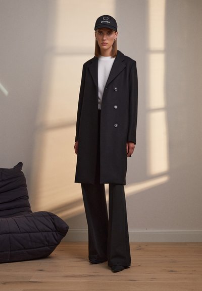 Black double-breasted coat with large buttons, worn over a white shirt, paired with black wide-leg pants and a black cap with a logo.