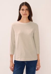 Frau mit langen braunen Haaren und Sommersprossen, die ein beiges, geripptes Langarmoberteil und blaue Jeans trägt und vor einem hellen Hintergrund steht.