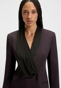 Blazer prugna con colletto a scialle in satin nero, vestibilità sartoriale e spalle strutturate. Presenta un unico gancio nascosto e tasche.