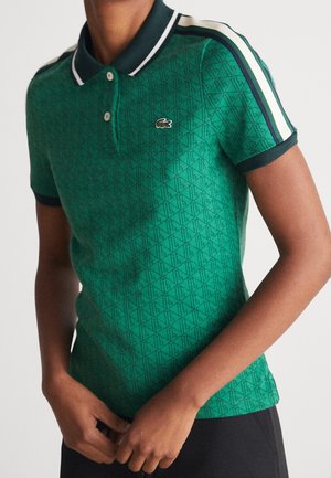 Zielona polo z wzorem, ciemnym kołnierzykiem i mankietami na rękawach, białe i granatowe paski na ramionach oraz małe logo krokodyla na piersi.