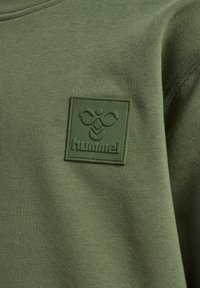 Zbliżenie na oliwkowo-zieloną tkaninę z naszywką w kształcie kwadratu, na której znajduje się stylizowane logo pszczoły i napis „hummel” w dopasowanym kolorze.