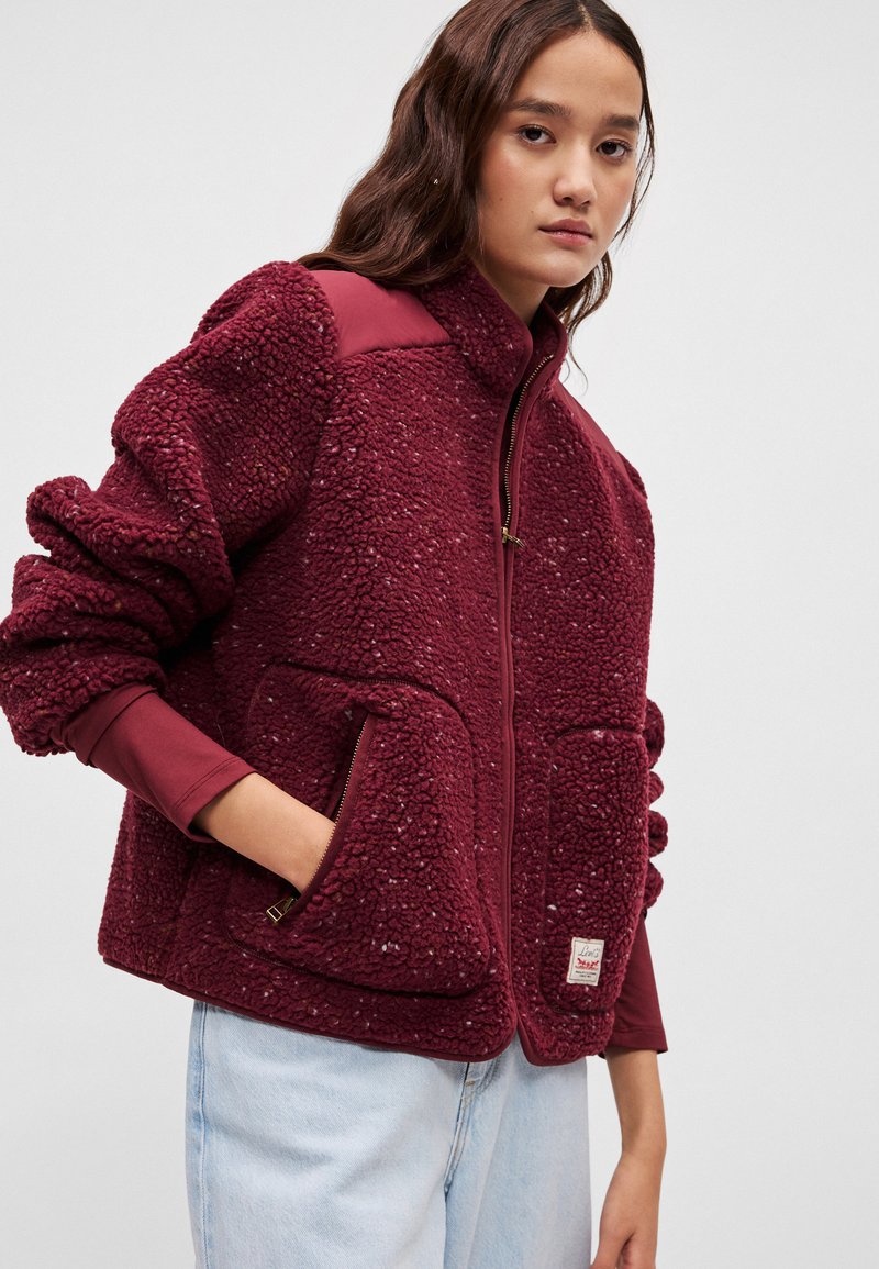 Burgundy fleece jacket featuring a zip front, two front pockets, and a textured finish. Long sleeves with ribbed cuffs.