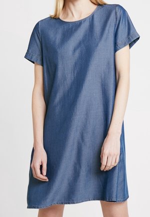 Kvinde iført en kortærmet, knælængde, denimblå shift kjole med rund halsudskæring, stående mod en ensfarvet baggrund.