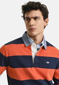 Camisa polo a rayas naranja y azul marino con cuello de mezclilla, que cuenta con una tapeta blanca y un pequeño logo en el pecho. Textura de tela suave.
