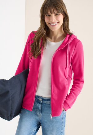 Femme souriante portant un sweat à capuche zippé rose vif, un t-shirt blanc et un jean bleu, tenant un grand sac bleu marine foncé sur une épaule.