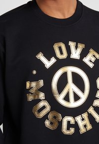 Svart sweatshirt med guldfärgat metalliskt tryck med texten "LOVE MOSCHINO" och en fredssymbol i mitten. Mjuk textur med ribbad halsringning.