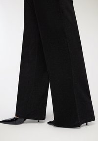 Czarne szerokie spodnie z teksturalnym, błyszczącym wykończeniem, zestawione z czarnymi, lakierowanymi szpilkami o stiletto.