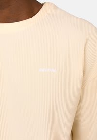 Maglietta a maniche lunghe color crema a coste con collo rotondo e sottile logo "CRYSTAL" ricamato in bianco sul lato sinistro.