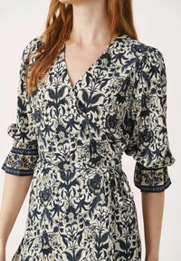 Robe portefeuille crème avec des motifs floraux bleus, dotée de manches courtes à volants, d'un décolleté en V profond et d'une taille nouée.