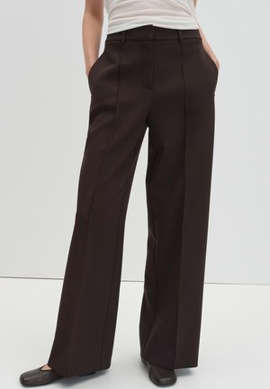 Frau trägt hoch taillierte, weit geschnittene dunkelbraune Hose und schwarze Schuhe, steht mit Händen in den Taschen vor schlichtem Hintergrund.