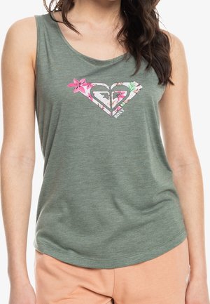 Femme portant un haut sans manches vert avec un logo floral en forme de cœur et un short pêche clair sur fond blanc.