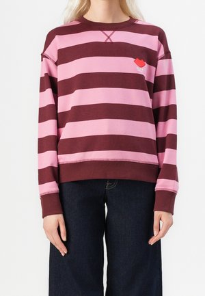 Kvinde iført en langærmet, pink og vinrød stribet sweater med mørke jeans, der viser torso og arme mod en ensartet baggrund.