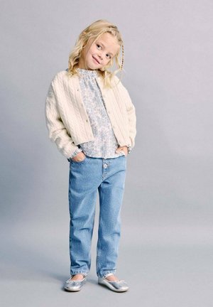 Fille blonde aux cheveux tressés, les mains dans les poches, portant un cardigan en maille blanc, un chemisier floral, un jean bleu et des ballerines argentées.