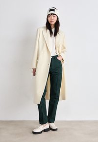 Dvojřadový béžový vlněný kabát, bílá košile, tmavě zelené elegantní kalhoty a krémové mokasíny s černými podrážkami, doplněné pletenou čelenkou.