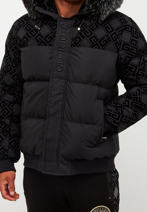 Homme portant une veste matelassée noire avec un motif géométrique sur les manches et la bordure de la capuche, avec le mot "GLORIOUS" sur le panneau avant.