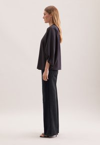 Mørkegrå oversized bluse med puffede ærmer, parret med højtaljede sorte denimjeans og sorte strophæle. Glat stoftekstur.
