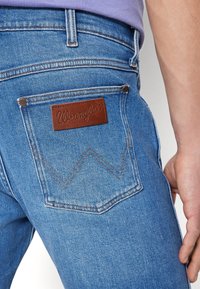 Närbild av en ficka på blå denimjeans med en brun läderlogopatch från Wrangler och en persons hand som vilar vid deras sida.