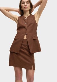 Gilet en lin marron avec quatre boutons et deux poches latérales, associé à une mini jupe marron avec une fente à l'avant. Texture lisse tout au long.