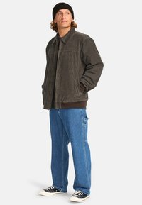 Giacca in velluto a coste grigia scura con colletto e zip, abbinata a jeans cargo blu e sneakers nere. Indossata con un berretto nero.