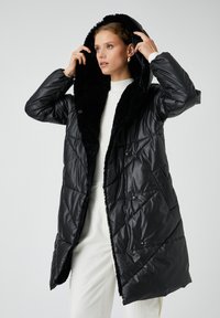 Vrouw in een zwart gewatteerd jack met een met bont gevoerde capuchon over een witte trui en broek, die de capuchon aanpast terwijl ze opzij kijkt.