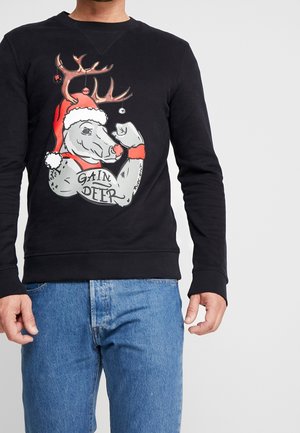 Uomo con felpa nera con grafica di testa di cervo muscoloso con cappello di Babbo Natale che si mostra un tatuaggio sul braccio con la scritta "Gain Deer", abbinata a jeans blu.