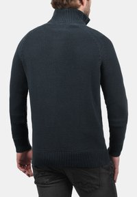 Jersey de punto azul oscuro con cuello alto, puños acanalados y dobladillo. Presenta una tela texturizada y un diseño ajustado, visible desde la parte trasera.