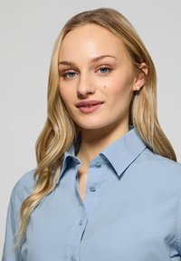 Ljusblå skjorta med krage, med en slät textur, en subtil logotyp och vanliga knäppningar.