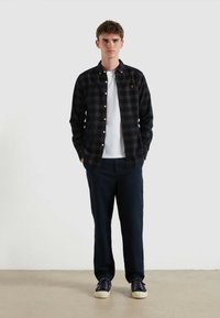 Camisa de cuadros oscuros sobre una camiseta blanca, combinada con pantalones oscuros y zapatillas navy. La camisa presenta un pequeño logo en el pecho.