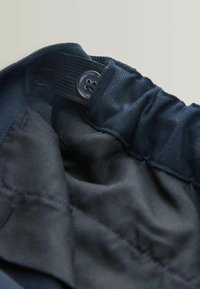 Indvendigt navyblåt stof med elastisk taljebånd; har en klar knap og glat tekstur. Syninger er synlige på sømmene, minimal detaljering.