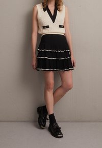 Pletená krémová vesta s černým límcem a dvěma předními kapsami; vrstvová černá skládací sukně s bílým lemem; masivní černé boty s detailem mašle.