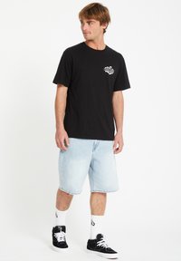 Schwarzes Baumwoll-T-Shirt mit einem Logo auf der linken Brust, kombiniert mit hellblauen Jeansshorts. Das Modell trägt schwarze Sneakers und weiße Socken.