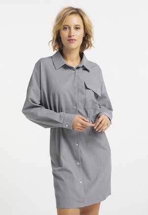 Junge Frau mit kurzen blonden Haaren, die ein lockeres graues Hemdkleid mit Brusttasche vor einem schlichten weißen Hintergrund trägt.