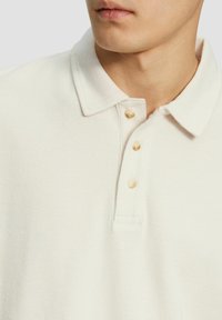 Weißes strukturiertes Polohemd mit einem Kragen, ausgestattet mit drei beigen Knöpfen und einer Brusttasche, die ein gewebtes Stoffmuster zeigt.