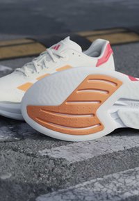 Chaussures de sport blanches et orange sur l'asphalte, une chaussure montrant une semelle orange texturée avec motifs antidérapants, lacets attachés sur l'autre chaussure.