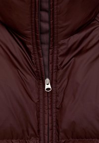 Burgundy puffer jacket featuring a central zipper with a silver pull tab, quilted texture, and contrasting black stitching.