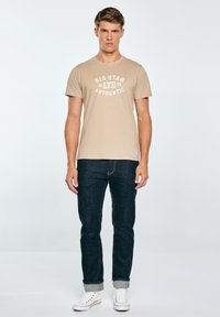 Beež puuvillane t-särk valge trükiga "BIG STAR 79 AUTHENTIC", kombineeritud tumeliste teksapükste ja valgete tennistega.