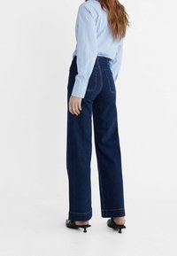 Mujer vestida con unos pantalones vaqueros de tiro alto y pierna ancha de color azul oscuro, una camisa de manga larga a rayas de color azul claro y unos zuecos negros de tacón bajo, de pie contra un fondo blanco.