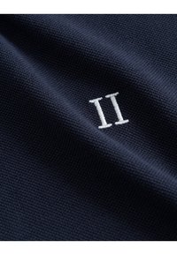 Tessuto navy con texture e un piccolo accento ricamato in bianco "II". La vista ravvicinata mette in evidenza la trama e i dettagli del materiale.