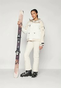 White and beige ski jacket with multiple pockets, beige ski pants, black ski boots, and a patterned ski with bindings, held upright.