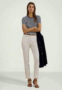 Mulher em pé com os braços cruzados, vestindo uma camisa listrada azul-marinho e branca, calças bejes, saltos castanhos e segurando uma jaqueta preta com um dos braços.