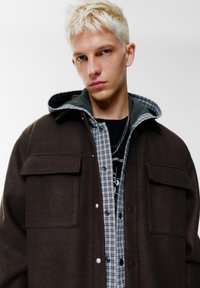 Giacca marrone in misto lana con chiusure a pressione, due tasche frontali, indossata sopra una camicia a quadri grigia e nera. Superfici dalla texture visibile.