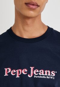 Tumsas zils kokvilnas T-krekls ar izteiktu sarkanu un baltu logotipu uz krūtīm, kas lasa "Pepe Jeans Portobello Rd W11", ar noapaļotu dekoltē.