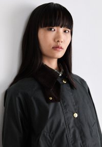 Dark green waterproof jacket featuring a brown corduroy collar, gold buttons, and a front pocket. The fabric has a smooth, matte texture.