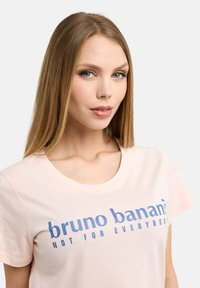 Rosa bomullströja med rund halsringning och blå tryckt text. Designen inkluderar varumärkesnamnet och en slogan i fet stil.