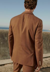 Brauner maßgeschneiderter Anzugjacke, glattes Material, strukturierter Schnitt, mit einem einzelnen Schlitz und hellen Knöpfen an den Ärmeln.