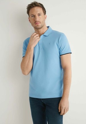 Mężczyzna z krótkimi włosami w jasnoniebieskiej koszulce polo i ciemnych dżinsach, stojący na tle gładkiej jasnej ściany.