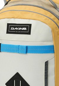 Mochila Dakine con tejido gris y amarillo, correa azul, parche de logo negro y acento triangular negro en la parte inferior.