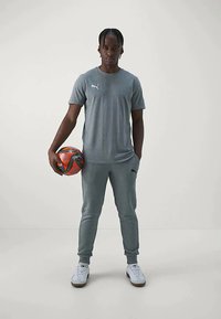 Grå sporty t-skjorte og matchende joggebukser, begge med en strukturert finish. Holder en oransje og svart fotball, iført hvite sneakers.