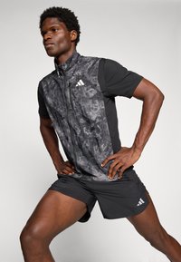 Sportlicher Mann dehnt seine Beine, trägt eine schwarze, gemusterte ärmellose Jacke und schwarze Shorts mit weißen Markenlogos, vor einem einfarbigen Hintergrund.