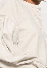 Vaalean beige corduroypaita, jossa pyöreä pääntie, napillinen nappilista ja runsaat hihat, joissa on olkapäiden kohdalla kerättyjä yksityiskohtia.